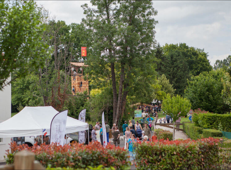 Des jeux en plein air sont installés dans un parc. L'un d'eux dépasse de la cime des arbres et une tente est visible au premier plan. Il y a beaucoup de monde dans le chemin