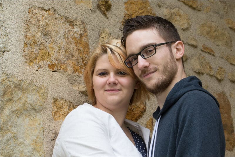 Deux amoureux posent tête contre tête devant un mur en pierre.