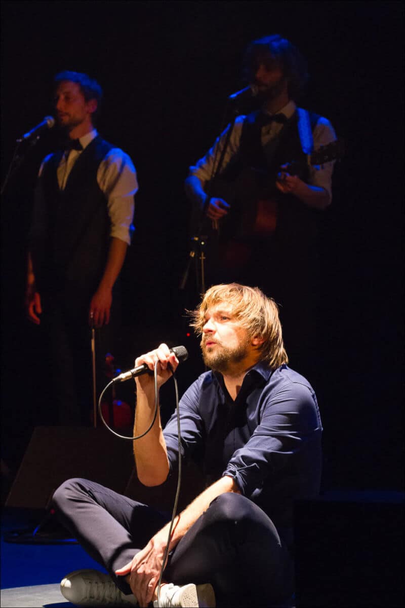 Le chanteur du groupe est assis sur la scéne, en pleine lumière, alors que deux musiciens sont derrière lui, dans l'ombre