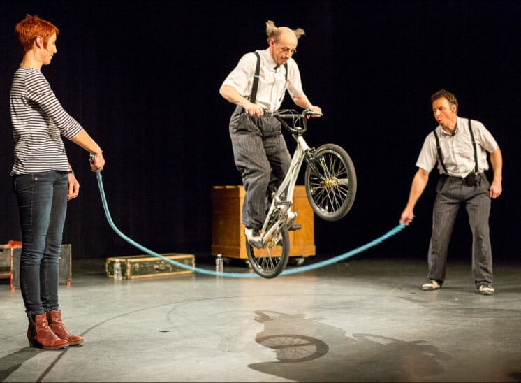 Deux personnes font tourner une corde à sauter alors qu'un autre artiste saute par dessus avec un vélo