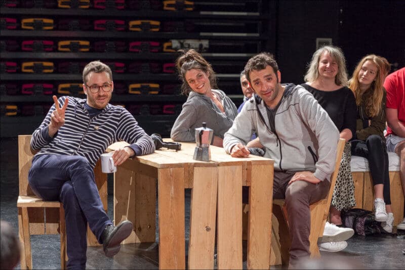 Les trois artistes sont assis autour d'une table en bois. L'un d'eux fait un signe de V avec ses doigts