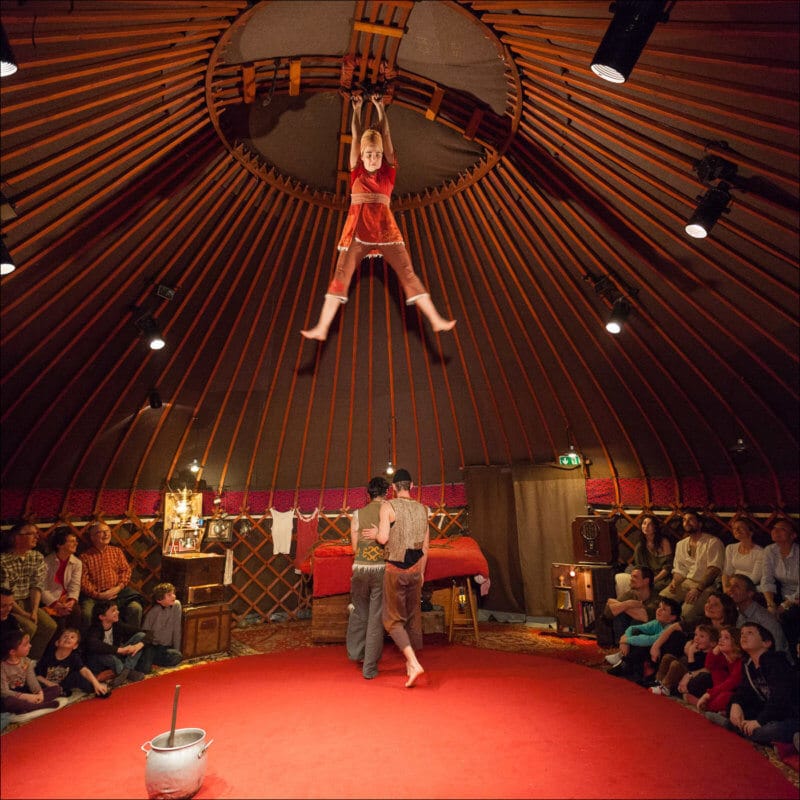 A l'intérieur d'un yourte, une femme est accrochée au plafond alors que deux homme discutement au dessous. Le public de ce spectacle est assis tout autour de la scène